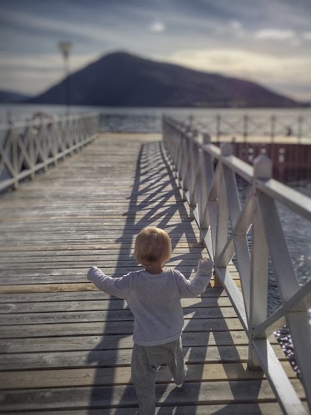 A toddler walking down a wooden dock on a sunny day with mountains in the distance.