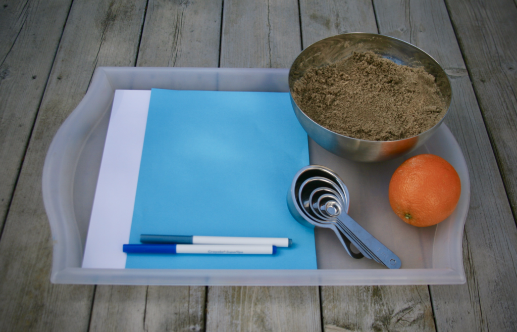 A plastic tray containing white paper, blue paper, blue pens, stainless steel measuring spoons, a stainless steel bowl full of sand and an orange on a wooden terrace