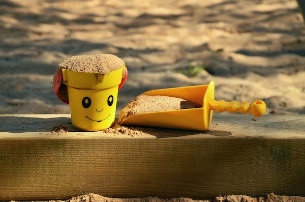Sand inside a yellow bucket with a smiley face and a yellow scoop on the wooden edge of a sand pit