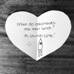 Science joke for kids: When do astronauts eat their lunch? At launch time! Handwritten on a heart-shaped note.