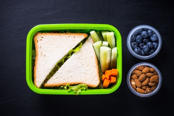 Green lunchbox with sandwiches and vegetables together with pots of blue berries and almonds on a dark table